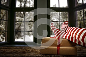 Woman legs in red and white christmas stockings and christmas presents on wooden table, snowy winter outside the window background
