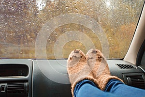 Woman legs in warm cute socks on car dashboard. Drinking warm tee on the way. Fall trip. Rain drops on windshield. Freedom travel