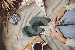 Woman legs in warm socks on soft blanket with cup of tea, christmas stars, golden lights, trees, candle and pillows, top view.