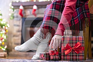 Woman legs in a winter socks covered plaid sitting and relaxation on armchair near fireplace and christmas tree pakking gift boxes