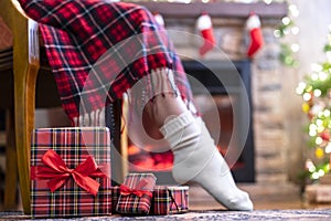 Woman legs in a winter socks covered plaid sitting and relaxation on armchair near fireplace and christmas tree pakking gift boxes