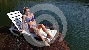 Woman lie on a sunbed in sunglasses and swimming suit. Girl rest on a flood wood underwater pier. The pavement is