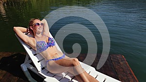 Woman lie on a sunbed in sunglasses and swimming suit. Girl rest on a flood wood underwater pier. The pavement is