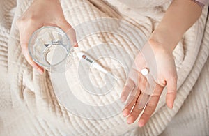 A woman lies in bed with a blanket and feels sick, holds a glass of water, pills and thermometer