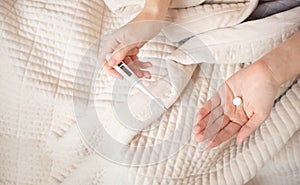 A woman lies in bed with a blanket and feels sick, holds a pills and thermometer in detail