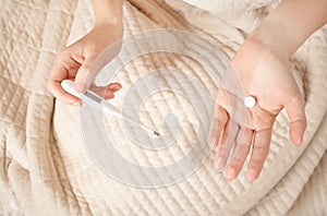 A woman lies in bed and feels sick. Female hands hold electronic thermometer and pills