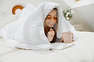 A woman lies on the bed under a blanket, smiling as she writes in her notebook