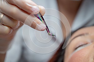 Woman lies on eyelash extension procedure in a beauty salon. Lashmaker holds tweezers with a bunch of artificial