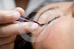 Woman lies on eyelash extension procedure in a beauty salon. Lashmaker holds tweezers with a bunch of artificial