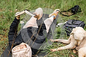 A woman lies on the grass and reads a book. A dog is resting nearby. Camping, relaxation