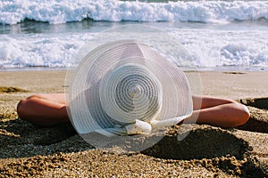 The woman lies on the sand against the background of the sea surf in a white hat with wide brim