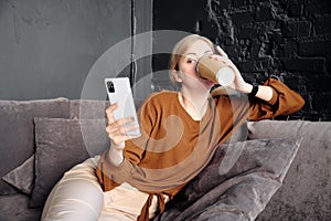 Woman lies on the sofa looks to the phone screen and drinks coffee