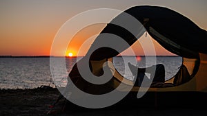 A woman lies in a tourist tent and enjoys the sunset by the river.