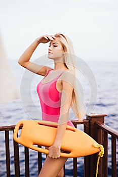 woman lifeguard with long blonde hair, standing in bikini with rescue board and looking on the sea