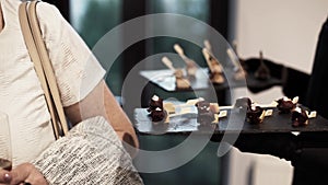 Woman in light clothes tries snacks on tray offered by waiter in black uniform.