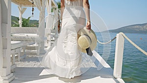 A woman in a light dress holding straw hat walking on a wooden pier, evoking holiday vibes and travel by the ocean.