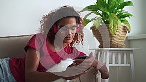 Woman listening to music and using smartphone while lying on couch in apartment room.