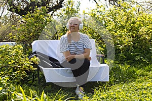 A woman listens to music with headphones while relaxing on a bench in a green garden.