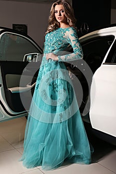 Woman with long blond hair wears luxurious dress,posing beside white car