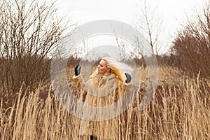 Woman with long blonde hair in fluffy coat standing in the tall dry grass, wind blowing and hair flying