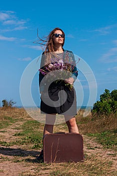 Woman with long brown hair in denim jacket, black skirt, vintage suitcase, flowers bouquet off-road. Lifestyle photo