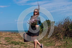 Woman with long brown hair in denim jacket, black skirt, vintage suitcase, flowers bouquet off-road. Lifestyle photo