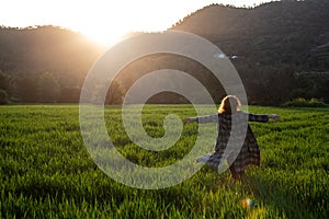 Woman with long coat and arms stretched enjoying sunset on the green meadow