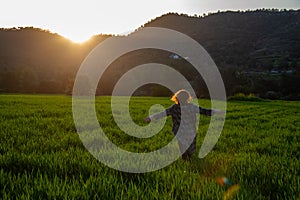 Woman with long coat and arms stretched enjoying sunset on the green meadow