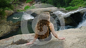 Woman with long curly hair sits on cliff edge, contemplates rushing waterfall below. Solo female traveler relaxes