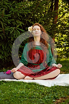 A woman with long curly red hair and closed eyes is sitting serenely on the grass in the lotus position. Meditations in nature