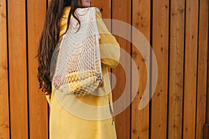 Woman with long dark hair wearing yellow raincoat holding string shopper bag with fruits standing back to the camera against