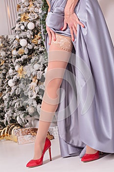 a woman in a long dress straightens her stockings on her legs in beautiful red stilettos near the New Year tree