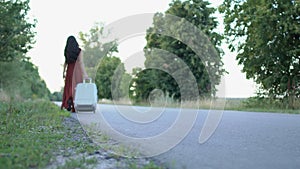 The woman in long dress walks with suitcase on the road