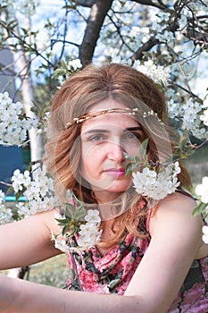 Woman with long hair in floral wreath looking at camera surrounded by blooming cherry branches