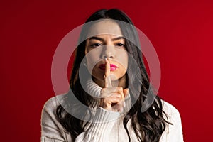 Woman with long hair holding finger on her lips over red background. Gesture of shhh, secret, silence. Close up.