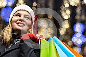 A woman with long hair and a Santa hat near the window of a city store with purchases in colorful, paper bags. New year`s shoppin