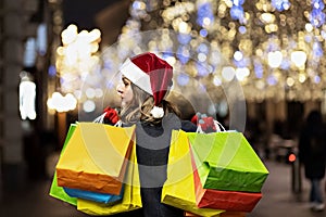 A woman with long hair and a Santa hat near the window of a city store with purchases in colorful, paper bags. New year`s shoppin