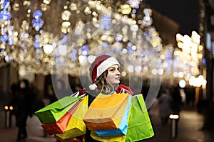A woman with long hair and a Santa hat near the window of a city store with purchases in colorful, paper bags. New year`s shoppin