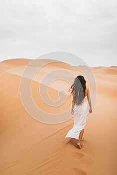 Woman with long hair in silk white dress walking in desert sand dunes, back view