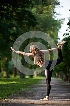 Woman with long hair standing in pose king of dance, Asan Natarajasana against greenery park under the sunny rays