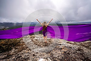 A woman with long hair is standing in a purple flowing dress with a flowing fabric. On the mountain against the