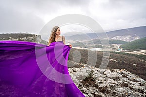 A woman with long hair is standing in a purple flowing dress with a flowing fabric. On the mountain against the