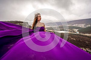 A woman with long hair is standing in a purple flowing dress with a flowing fabric. On the mountain against the