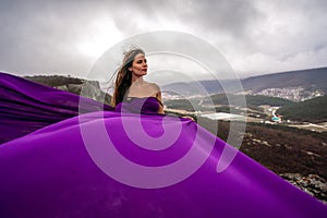 A woman with long hair is standing in a purple flowing dress with a flowing fabric. On the mountain against the