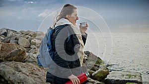 Woman with long hair, standing on the rocks and watching the winter waves while drinking hot tea. A great addition to travel or