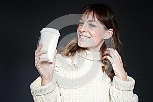 Woman with long hair with takeaway disposable cup of coffee in hands, wearing white winter sweater, dark grey background.