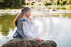 a woman with long hair in a vintage historical blue dress sits by the river 1