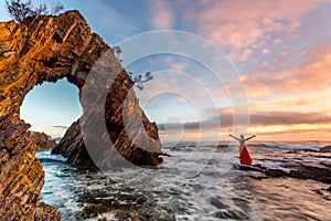 Woman in long red dress standing in ocean waves by a sea arch