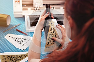 woman with long red hair and painted nails aligns christmas ornament sticker on a wine bottle.