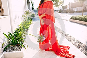 Woman in a long red translucent dress stands on the street next to a blooming flower bed against the backdrop of a house